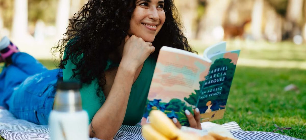Smiling woman reading a book at a picnic in a sunny park setting.