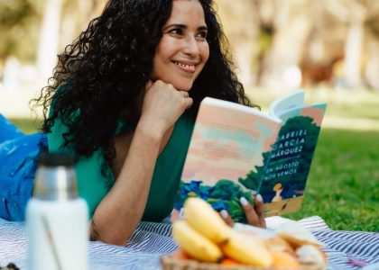 Smiling woman reading a book at a picnic in a sunny park setting.